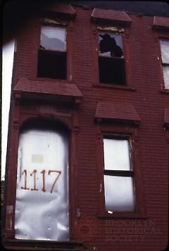 [Building facade with missing or broken windows and street number written on door]