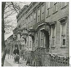 [Woman pushing baby carriage before row houses]